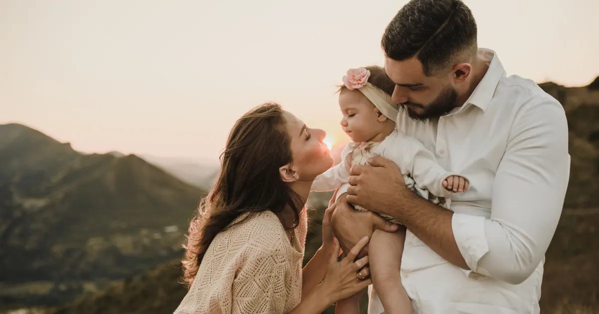 Parents holding a baby together on a hillside at sunset