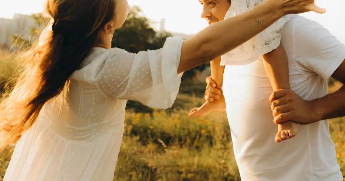 A family backlit by low sun in a meadow, a child riding on a parent's shoulders