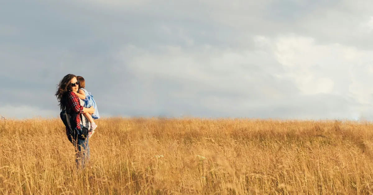 A parent carrying a small child, standing alone in a wide field of dry grass under a broad sky